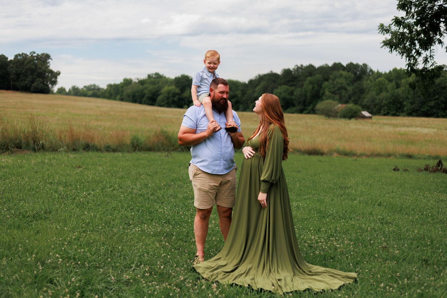 Family maternity photo with woman in flowing green gown, bearded man, and toddler on shoulders in a lush field.
