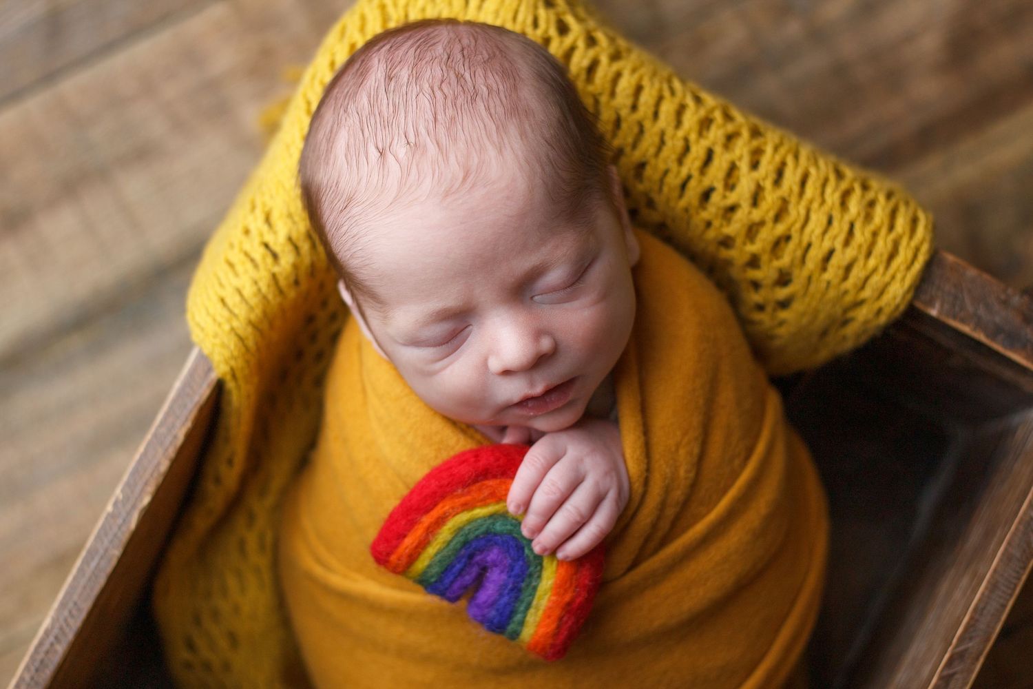 Sleeping newborn wrapped in a mustard yellow wrap holding a small rainbow prop during a newborn photography session in Dalton, Georgia.