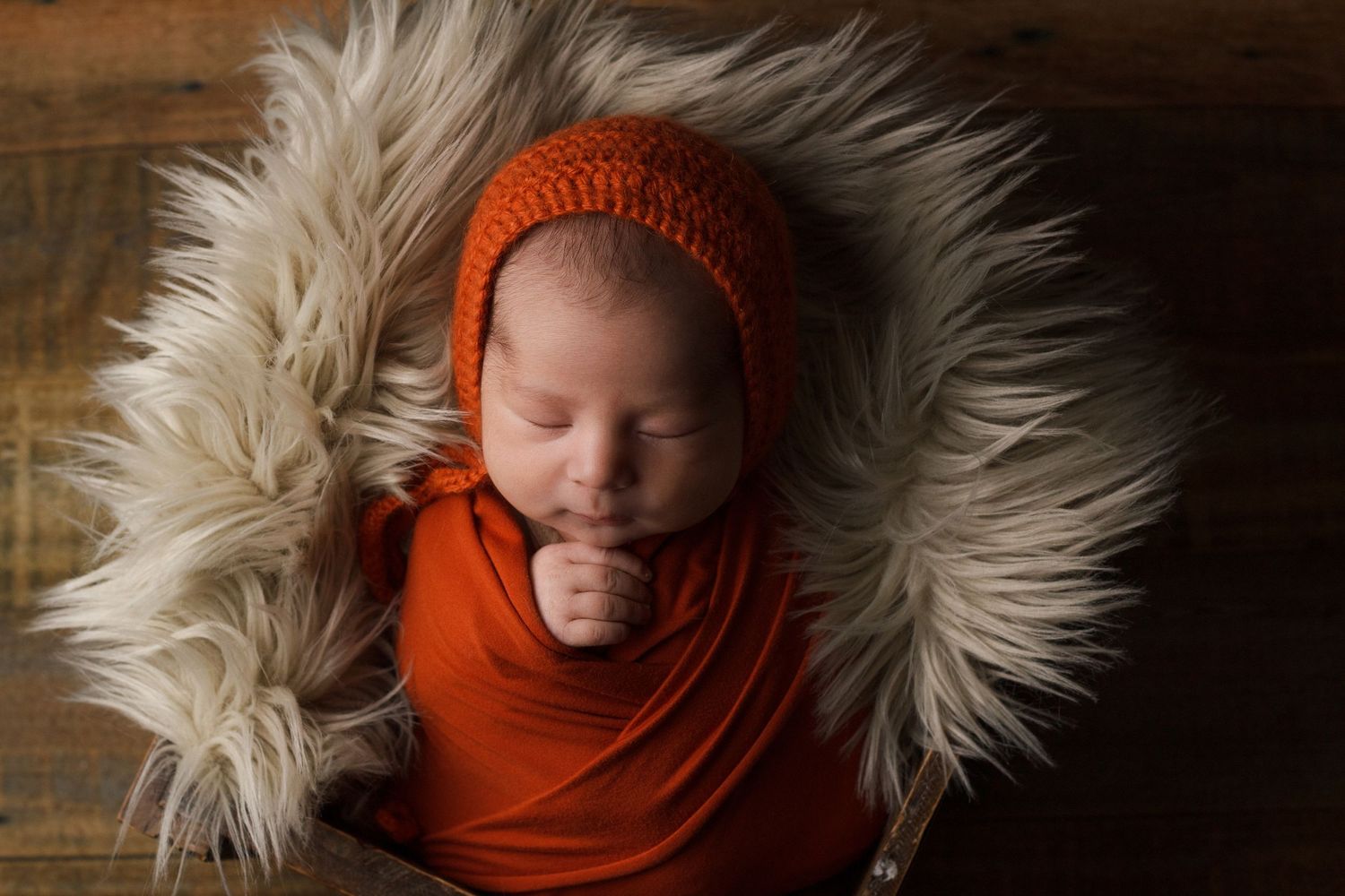 Newborn baby wrapped in a warm rust-colored wrap with matching bonnet sleeping on a grey fur layer during newborn session.