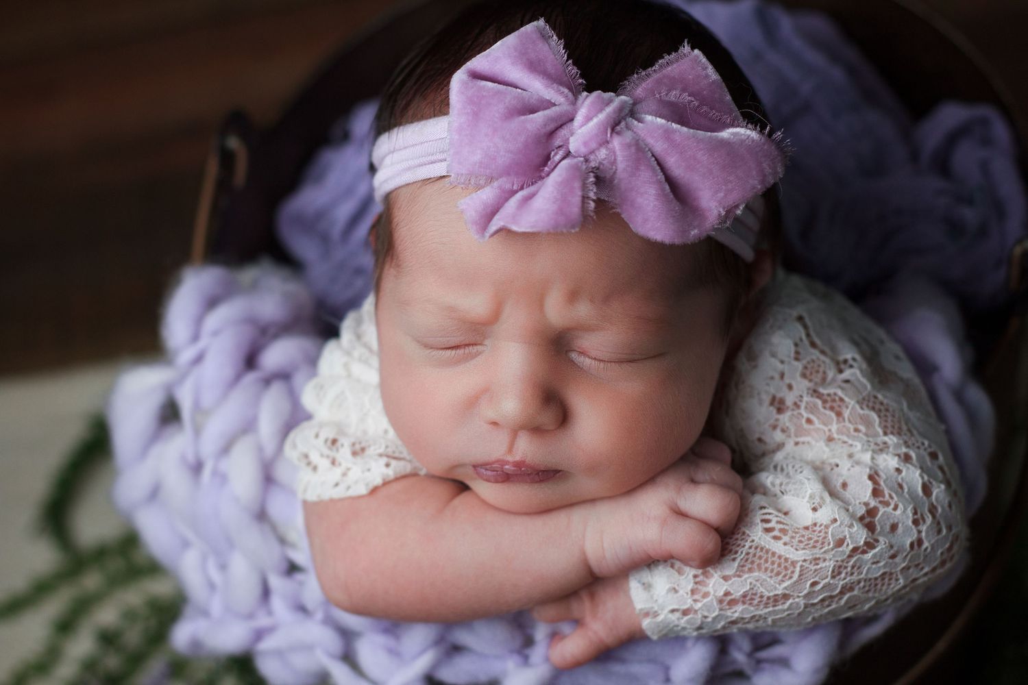 Baby girl  dressed in white lace and a purple bow headband resting her head on her arm during a newborn session in Dalton Georgia