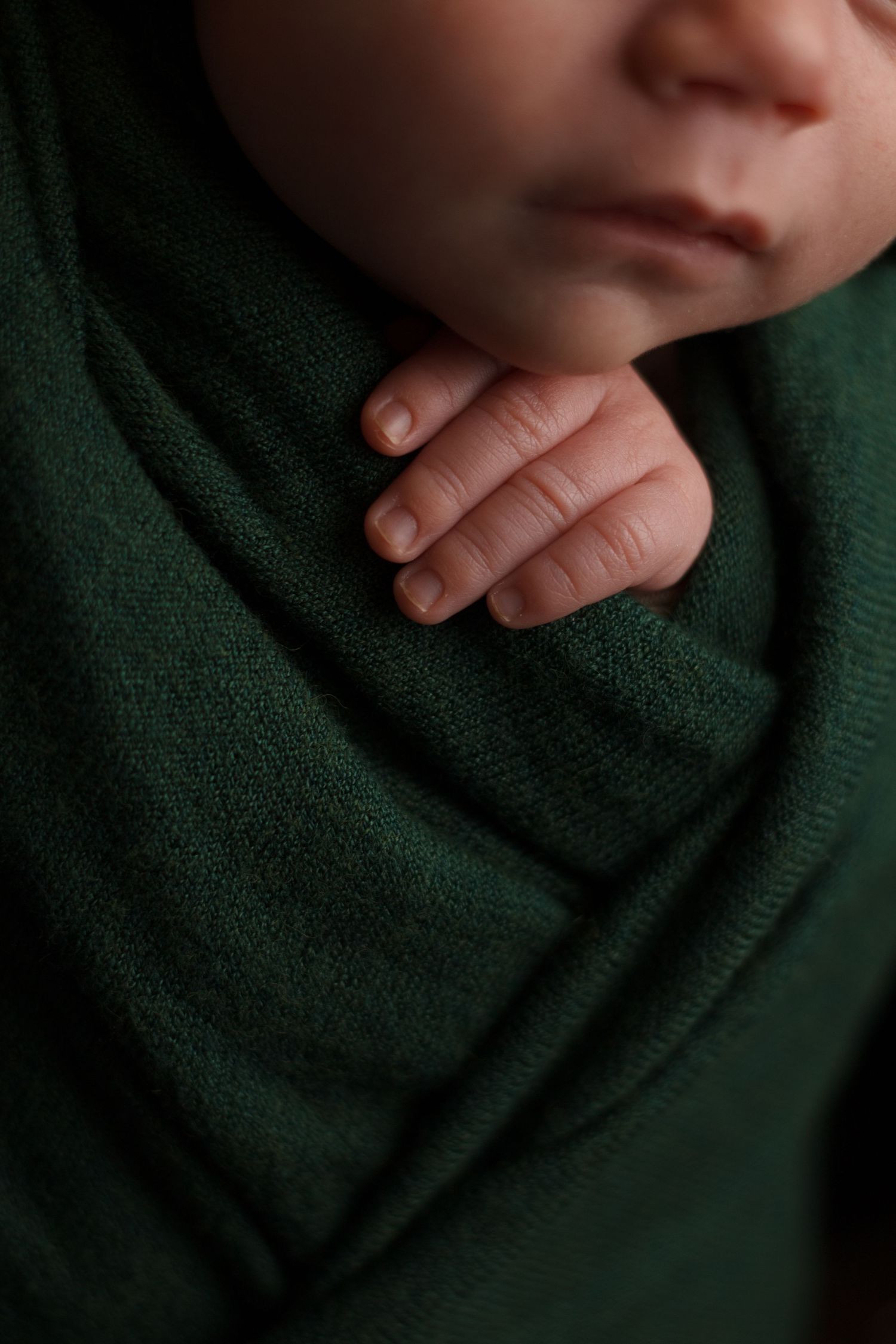 Close up of newborn baby wrapped in deep green resting peacefully with his hand out in a wooden prop during a newborn photography session.