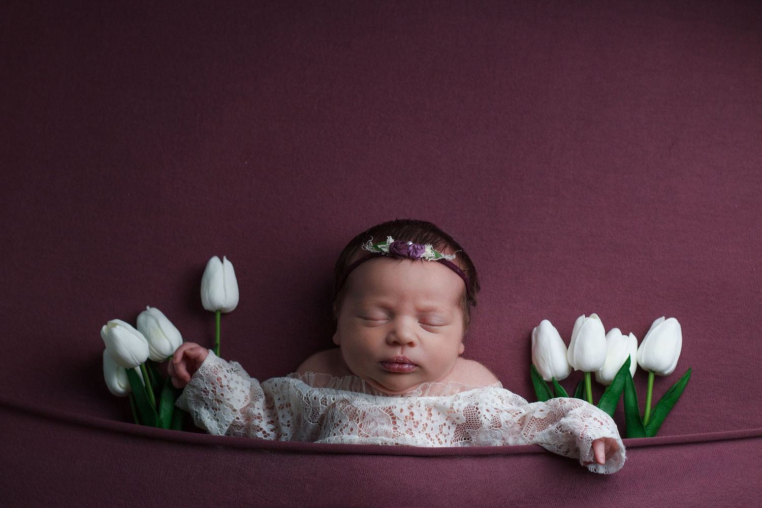 Newborn baby girl in white outfit laying on purple backdrop with with tulips tucked beside her on each side in Dalton Ga.