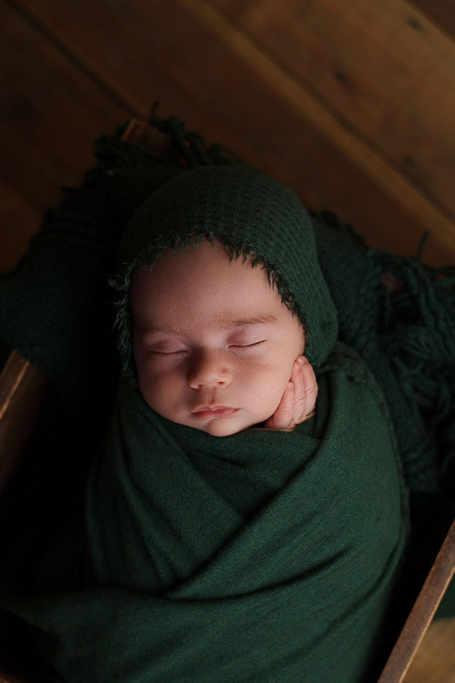 Newborn baby wrapped in deep green fabric resting peacefully with hand on cheek in a wooden prop during a newborn photography session.