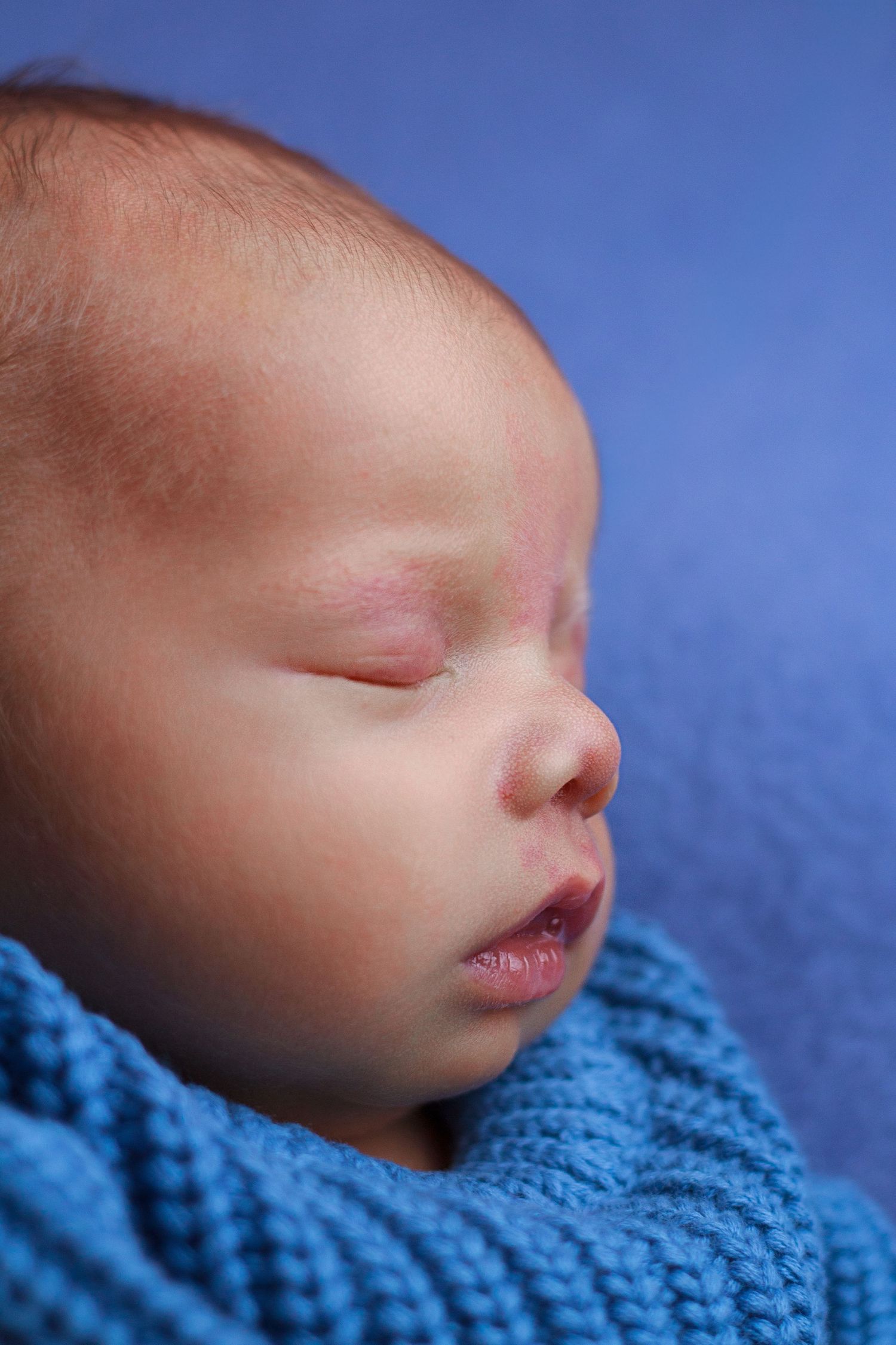 Close-up side profile of a newborn baby boy sleeping against a soft blue background.