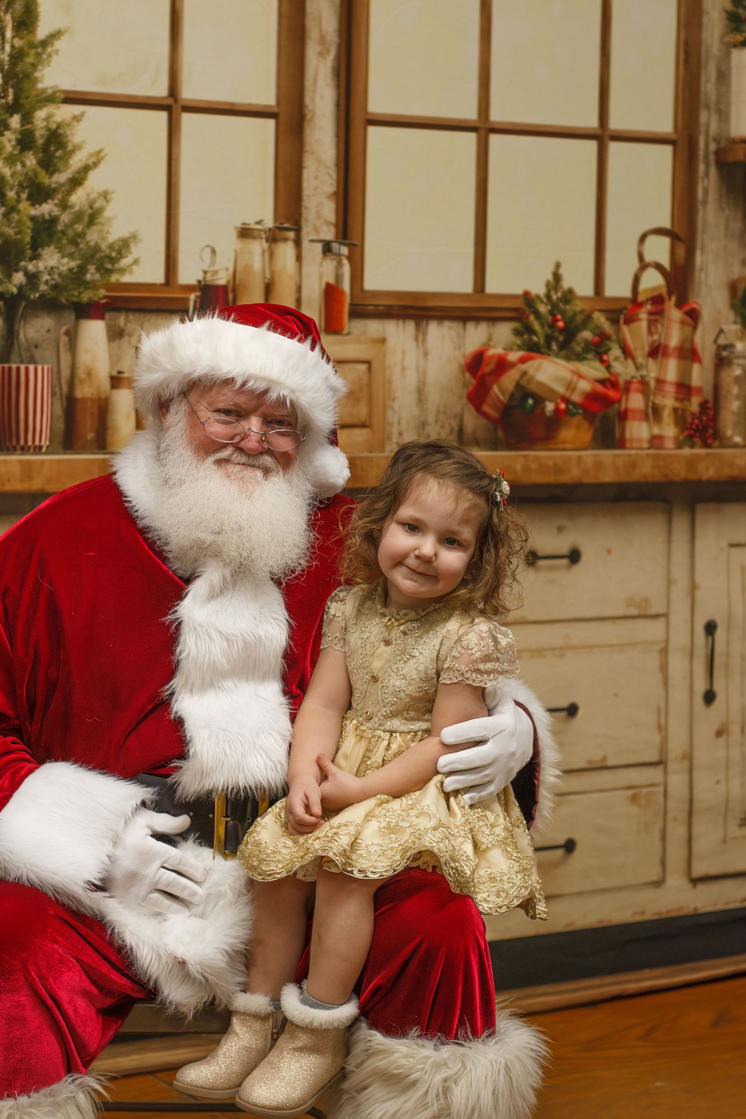 Smiling little girl in gold dress sits on Santa Claus lap in Christmas kitchen setting.