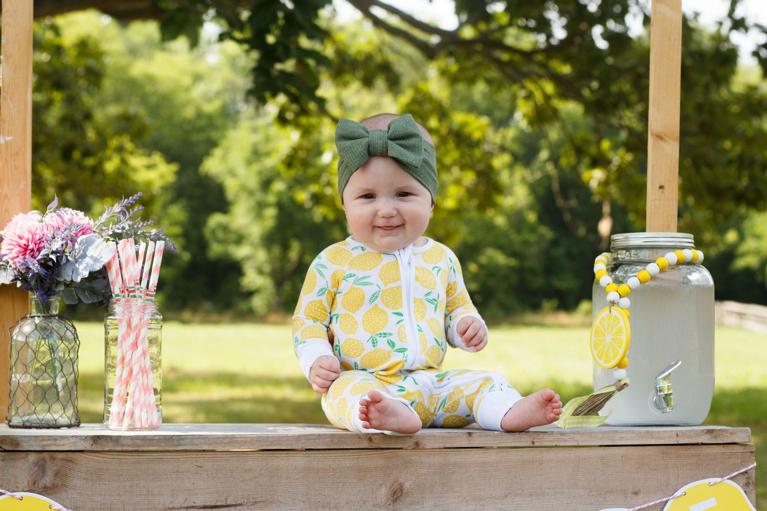 Baby girl dressed in a lemon sleeper sitting on a lemonade stand between flowers and lemonade during a Tunnel hill Georgia Milestone Session