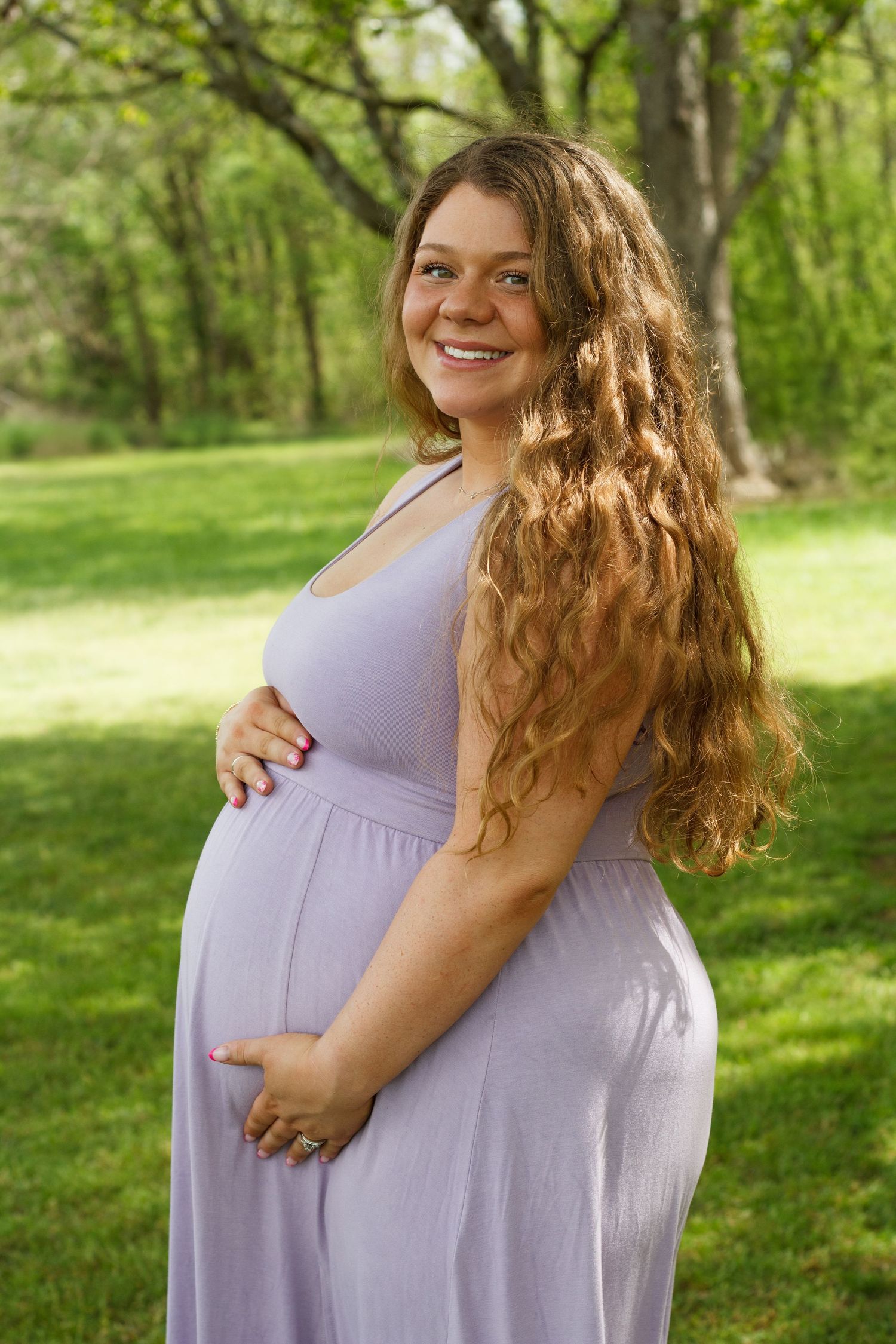 Smiling pregnant woman in lavender dress cradles baby bump outdoors in lush green park.
