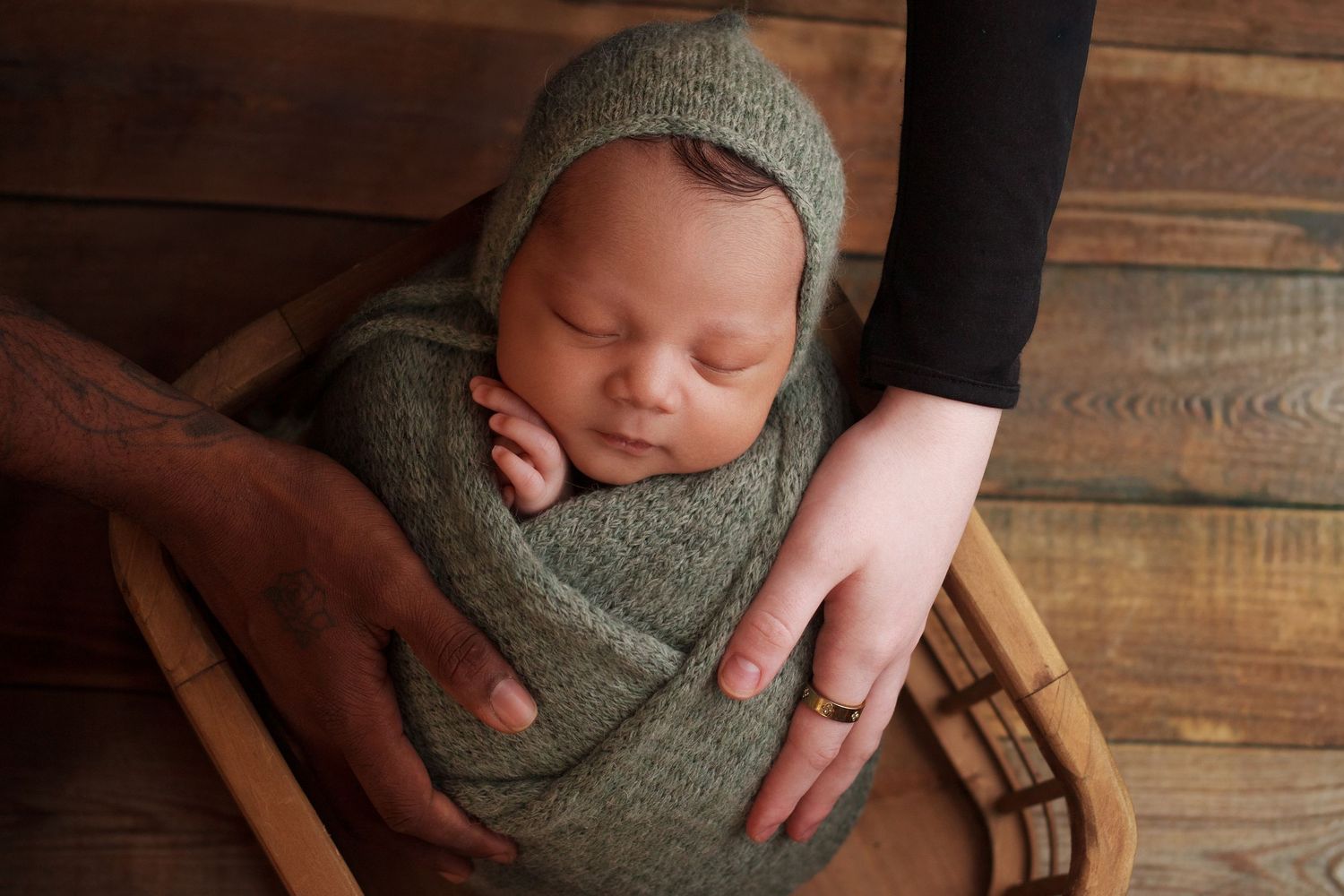 Baby boy wrapped in a sage green knit wrap surrounded by mom and dads hands in a wooden basket during his newborn session in Ringgold Georgi
