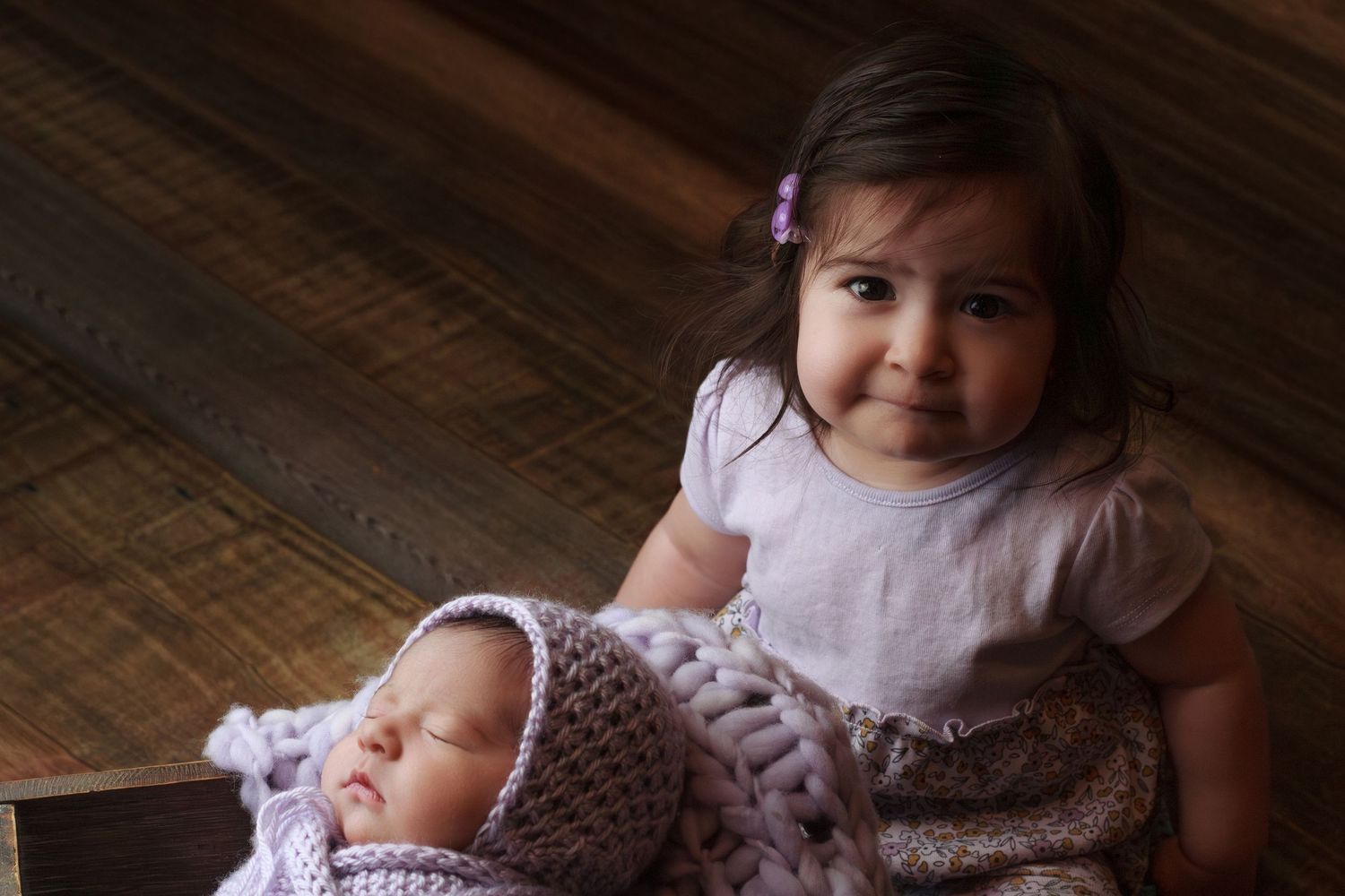 Toddler girl sitting on a wooden floor sitting next to her newborn sibling during a family session.