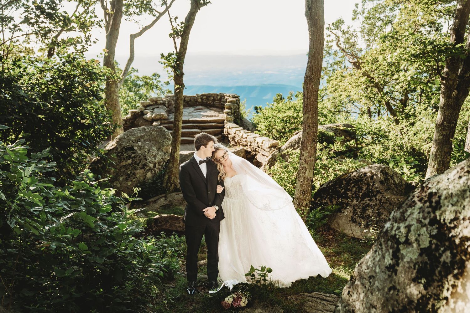 Bride and groom share a romantic kiss surrounded by lush forest trees and rocky terrain with scenic mountain views.