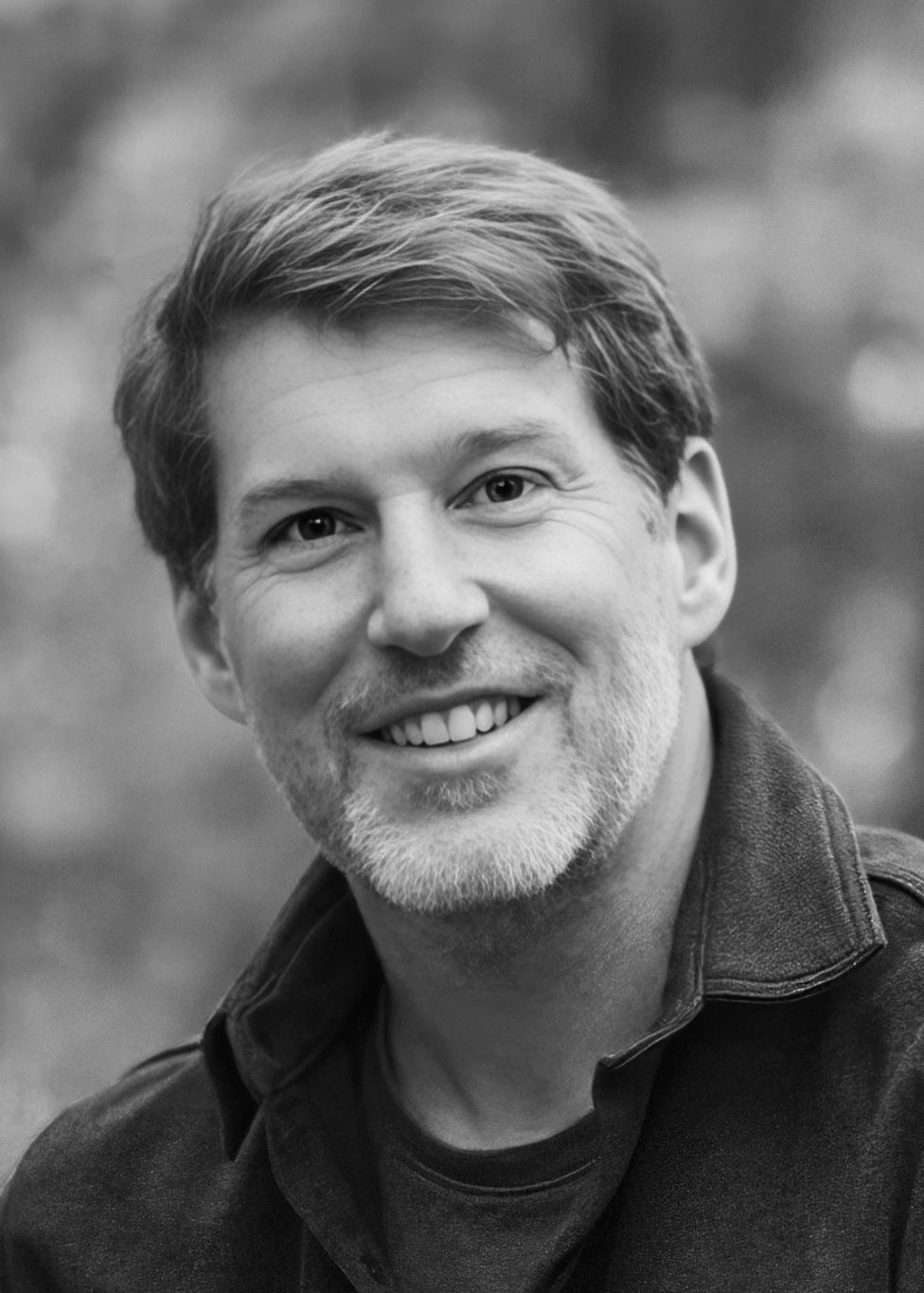 Black and white professional headshot of someone wearing a dark jacket and smiling warmly at the camera.
