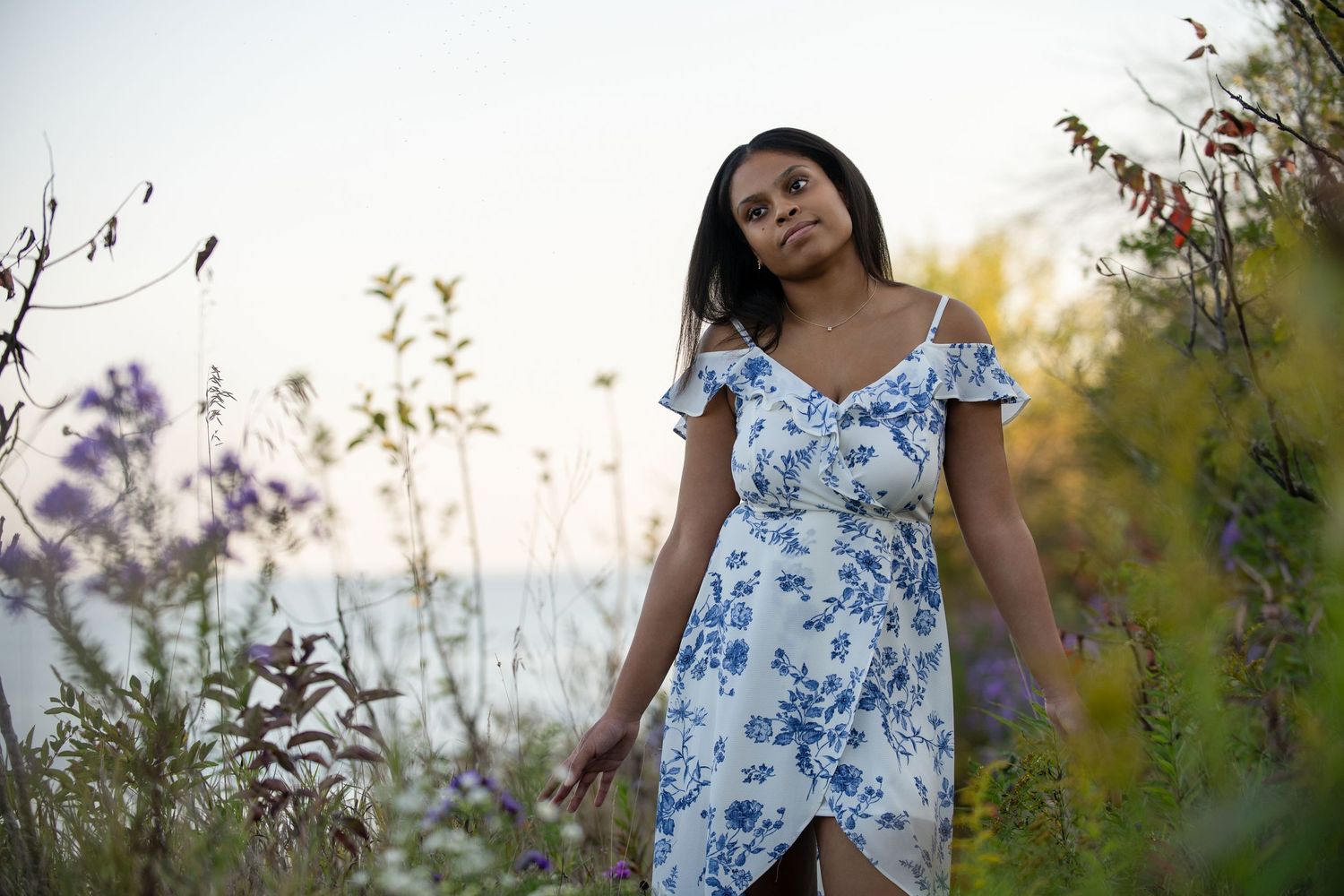 Young woman in white floral dress standing among wildflowers near water at dusk.