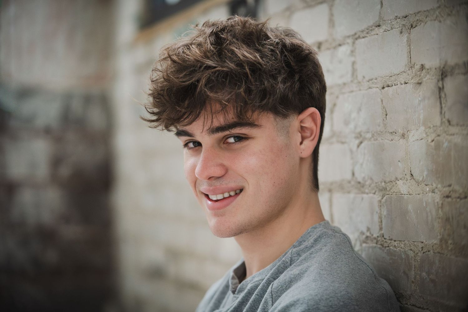 Smiling teenage boy with curly brown hair leaning against a white brick wall wearing a gray t-shirt.