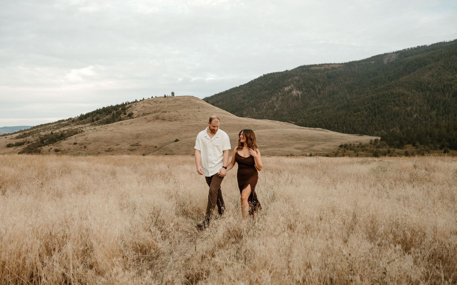 couple walking and laughing together in tall yellow grass with Vernon bc's rolling hills and big blue beautiful sky in the background