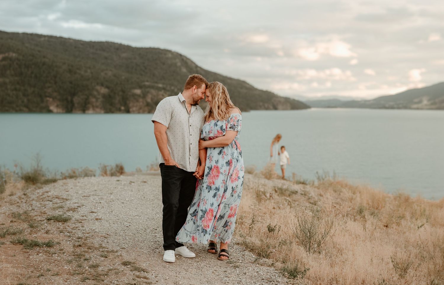 Mom and dad embracing with children playing behind them, kalamalka lake okanagan bc is the beautiful backdrop where these family photos were