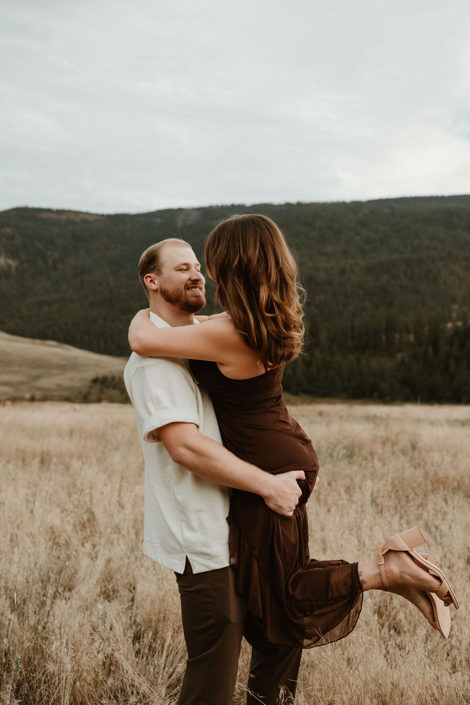 candid engagement photo of couple dancing in a grassy field with mountains in the background during Vernon bc engagement session