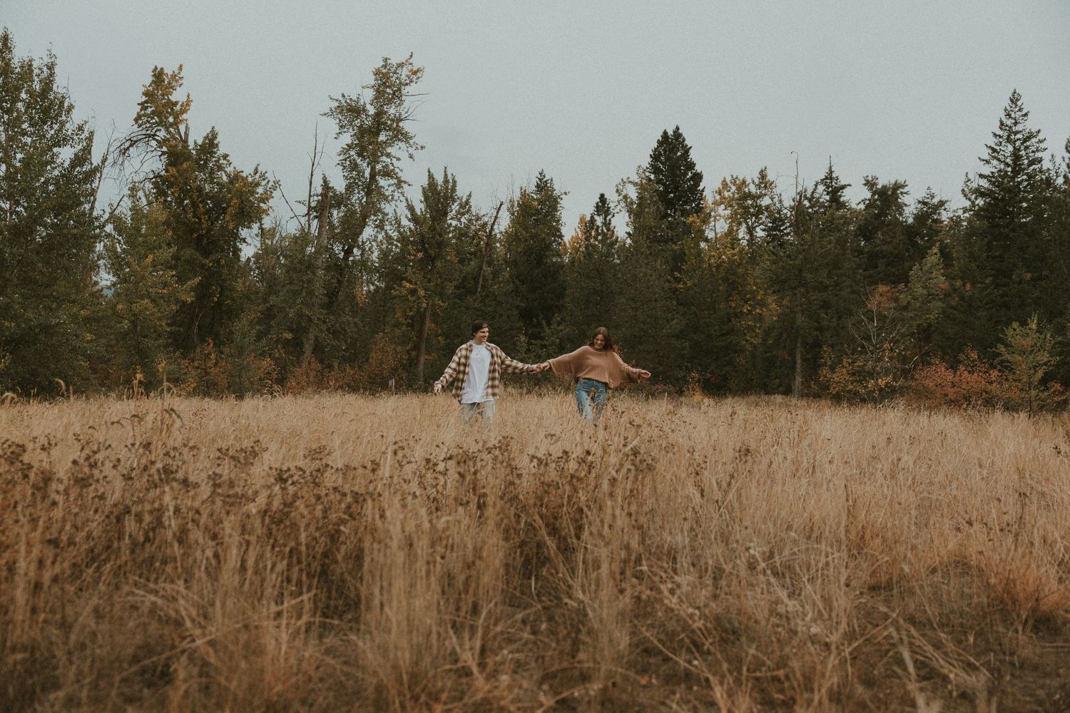 couple walking hand in hand through tall grass during moody okanagan engagement session