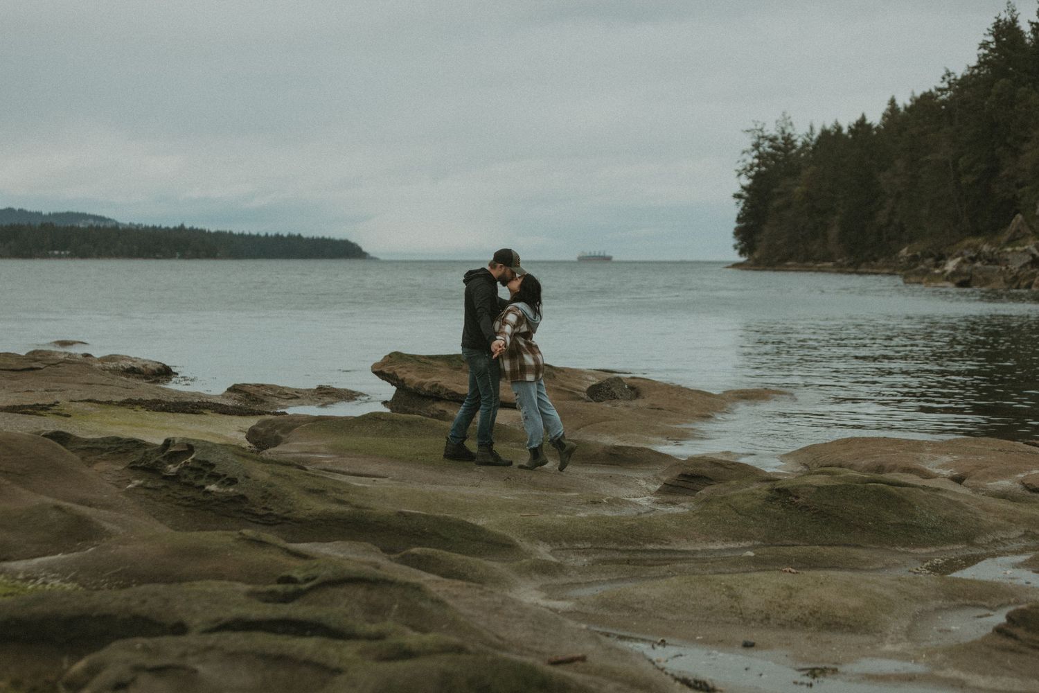couple standing on a rocky beach kissing with the tree lined Pacific Ocean in the background (nanoose bc)