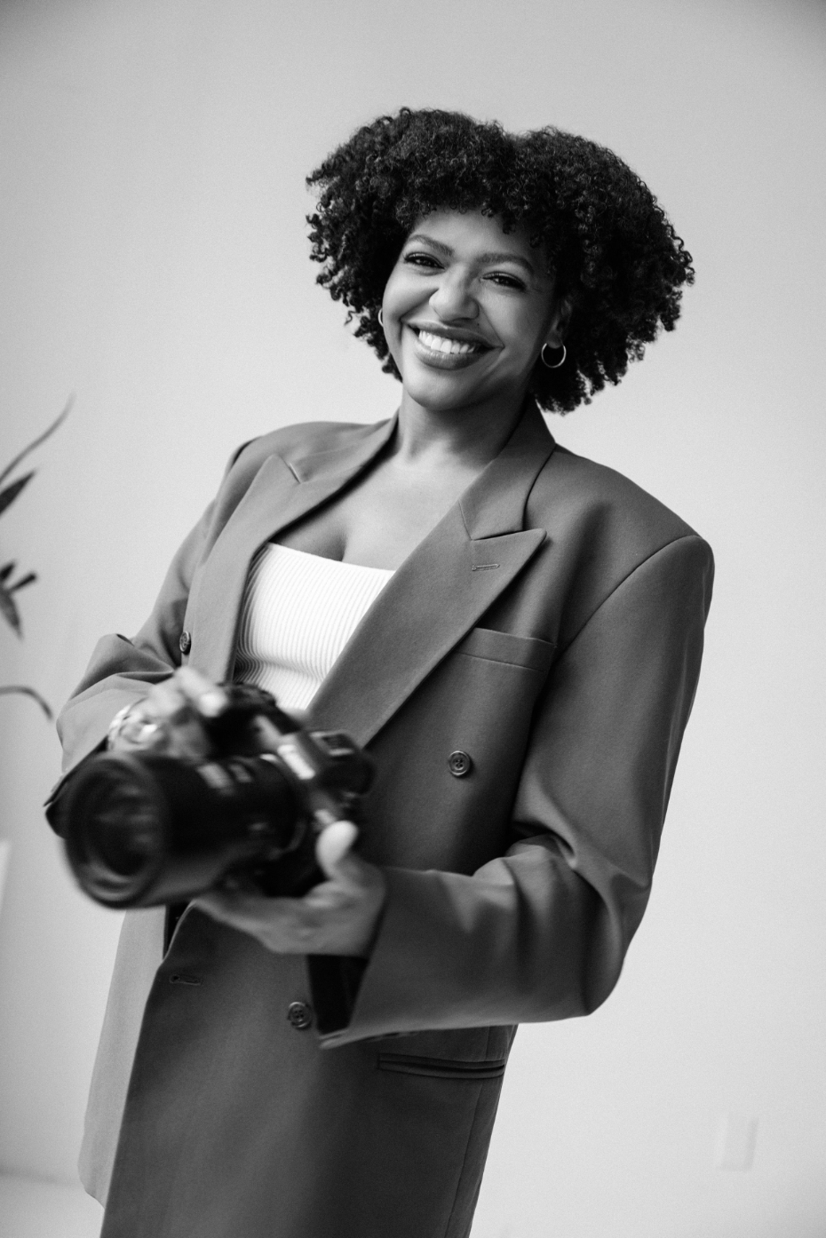 Confident woman with natural afro holds DSLR camera in elegant blazer in monochrome portrait.