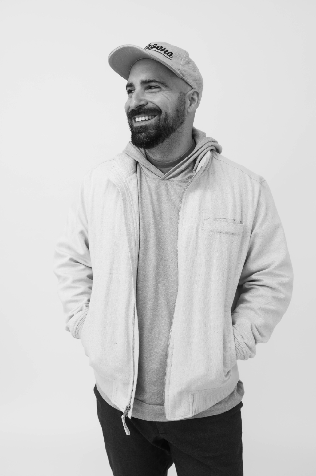 Bearded man in baseball cap and layered casual jacket smiles warmly in black and white photo.