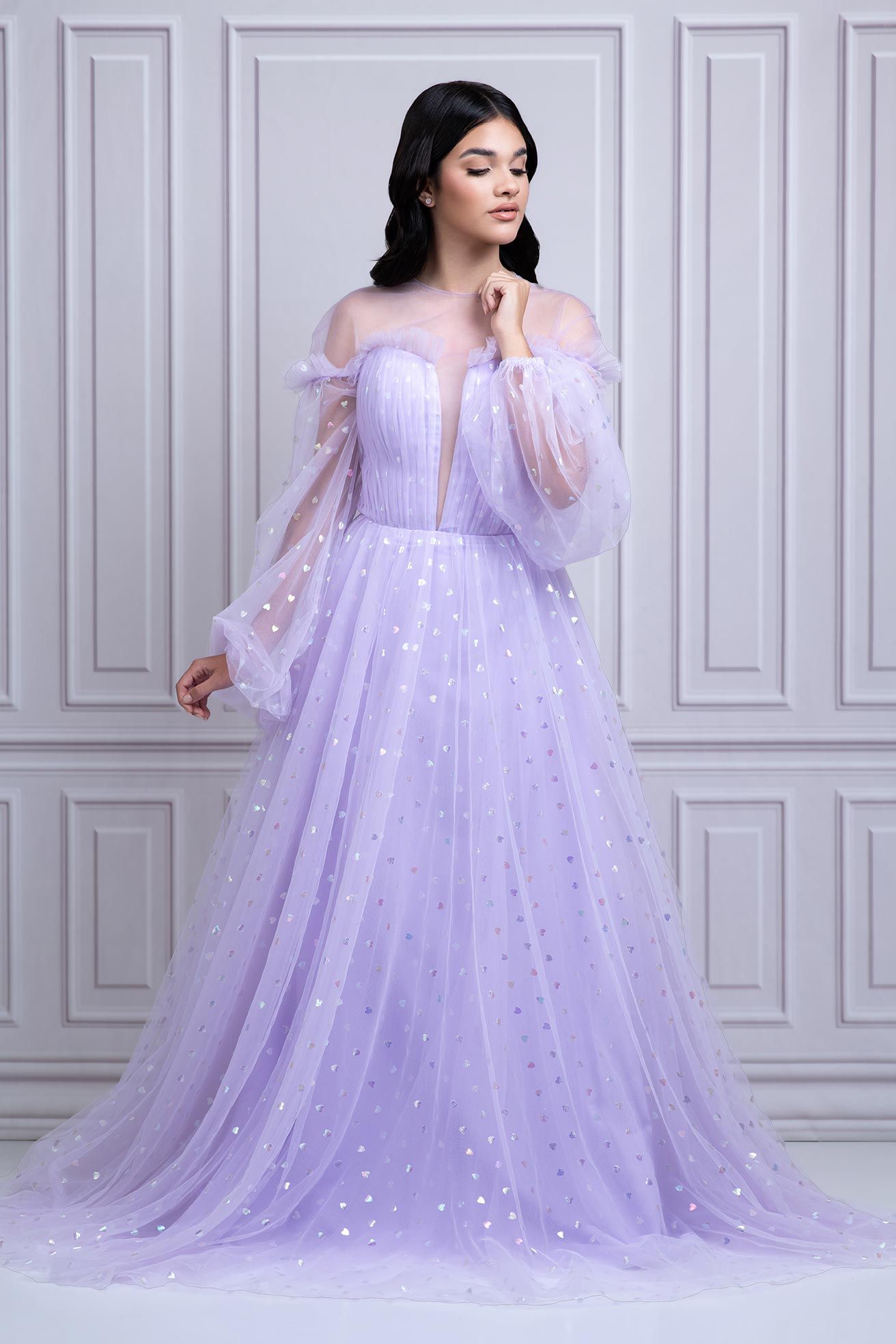 Ethereal lavender tulle gown with off-shoulder sleeves and sparkly star details flows elegantly against white paneled wall.