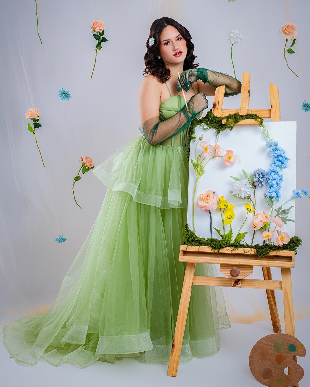 Artist in flowing sage green tulle gown stands beside an easel with floral painting against a wall with floating roses.