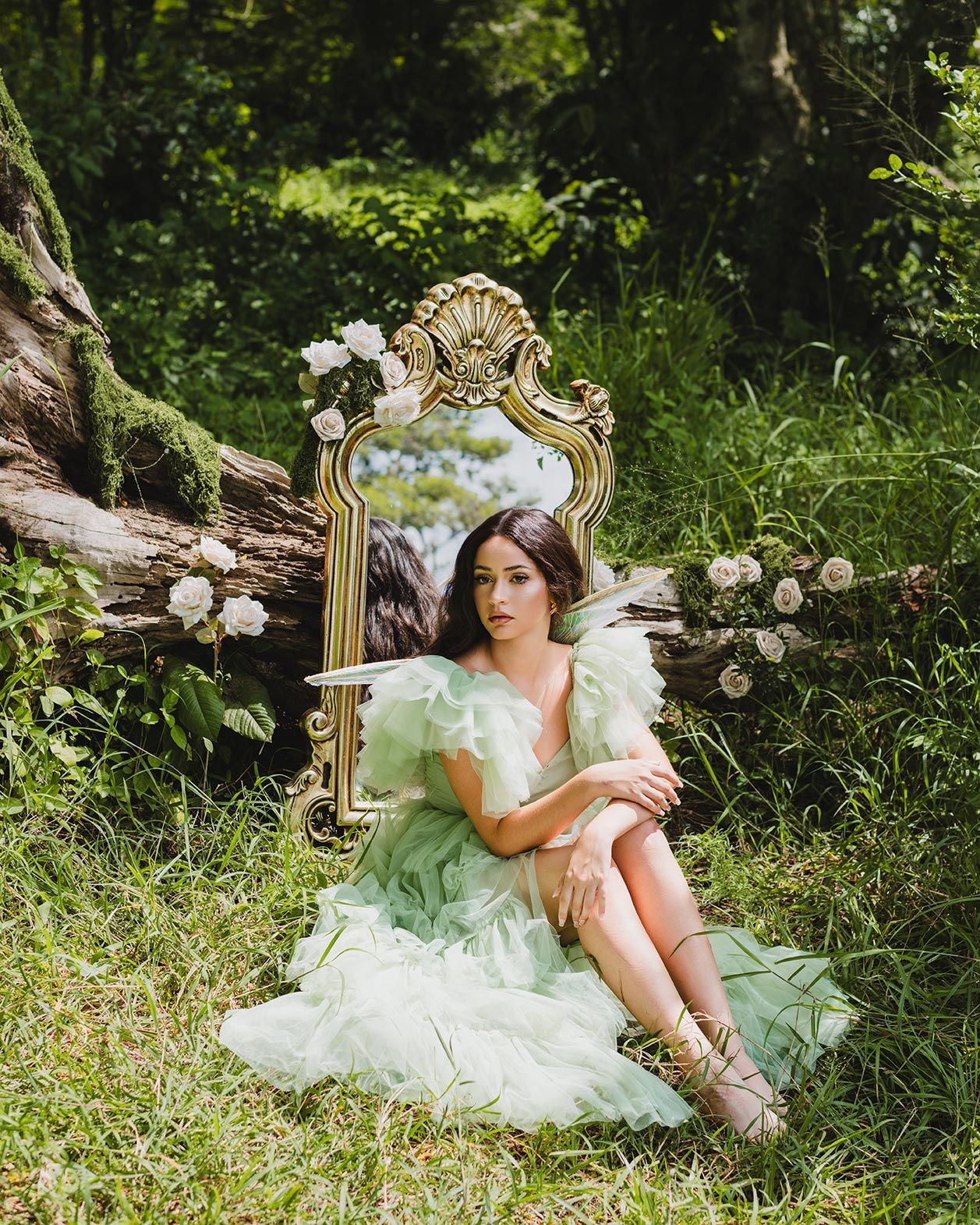A vintage ornate mirror rests against a fallen log surrounded by white roses and lush green forest scenery.