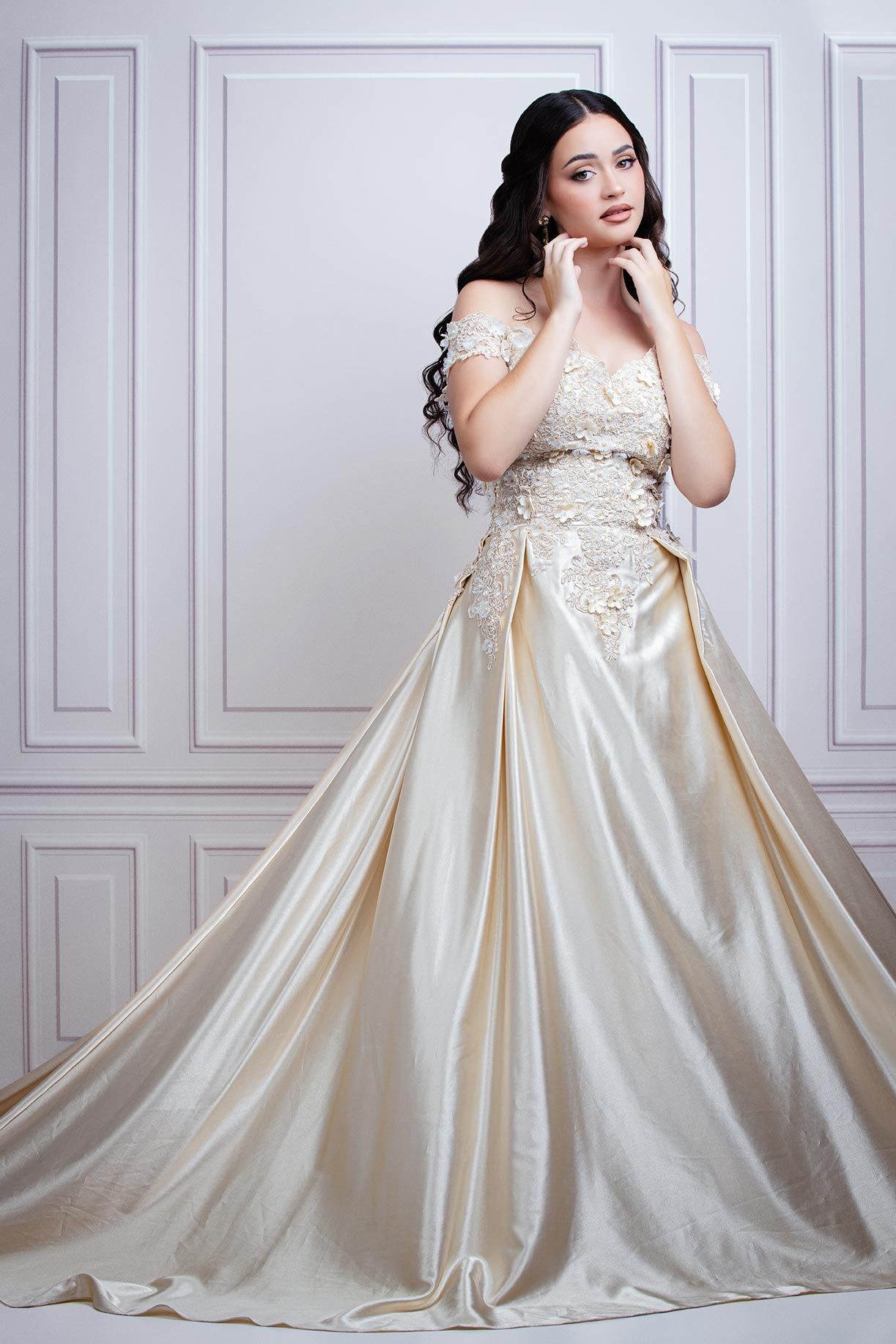 Elegant ivory satin wedding gown with lace bodice and off-shoulder design photographed against white paneled wall.