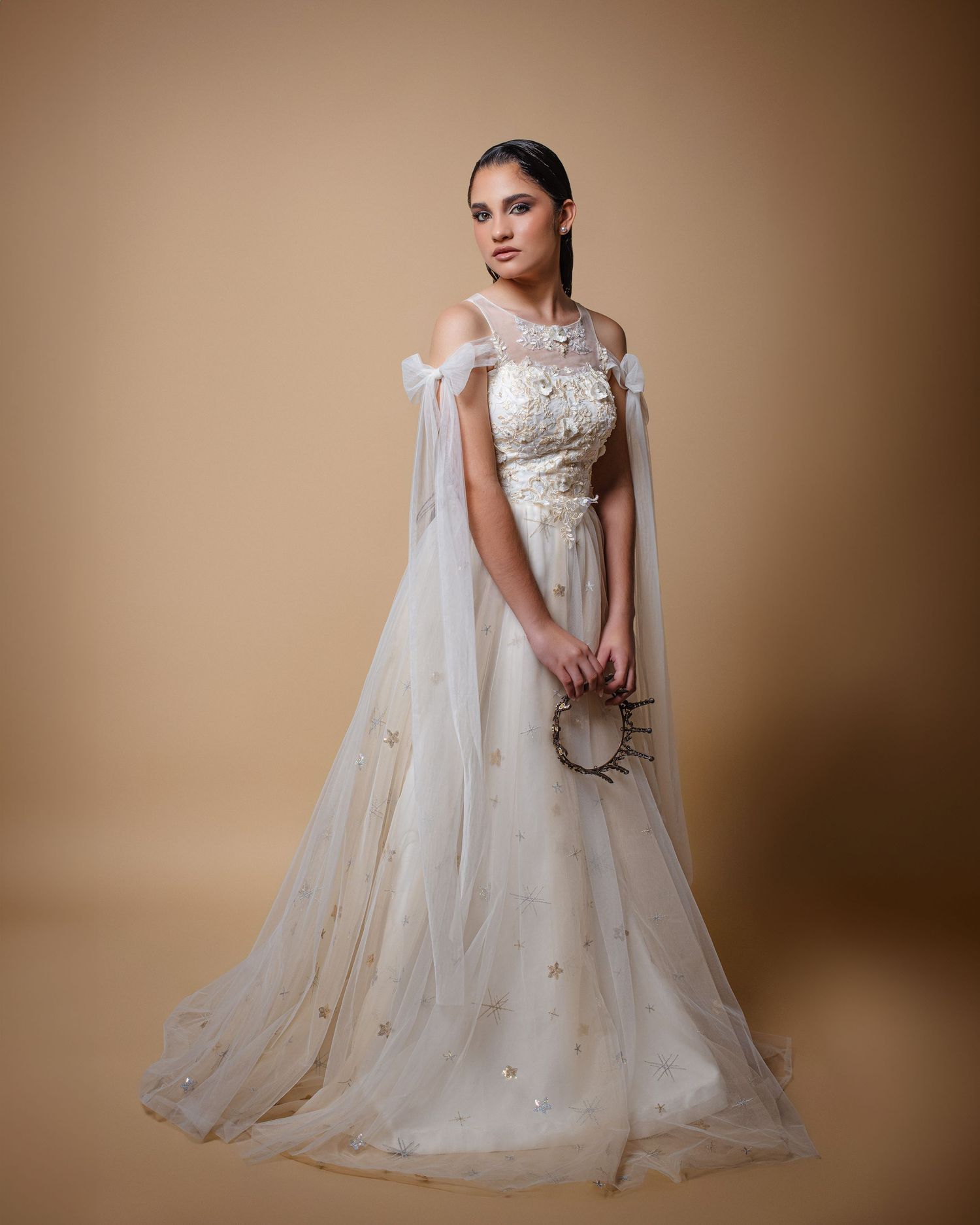Elegant off-shoulder wedding gown in white tulle with sheer cape, floral appliques and beaded details against beige backdrop.