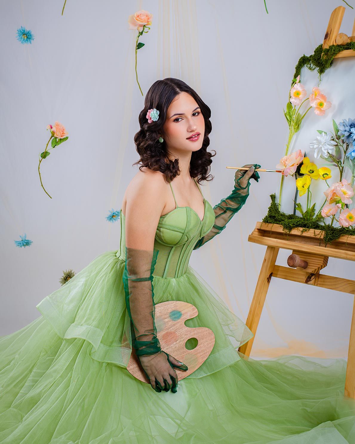 Whimsical art-themed photoshoot with green tulle ballgown, painter's palette, and floral decorations against studio backdrop.