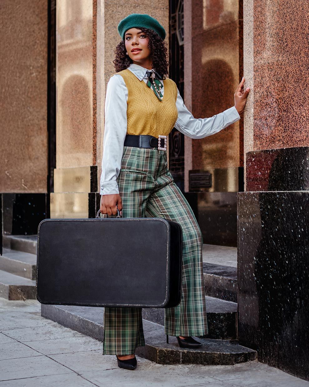 Stylishly dressed person in vintage-inspired outfit with plaid pants, yellow vest, and beret carries retro suitcase on city steps.