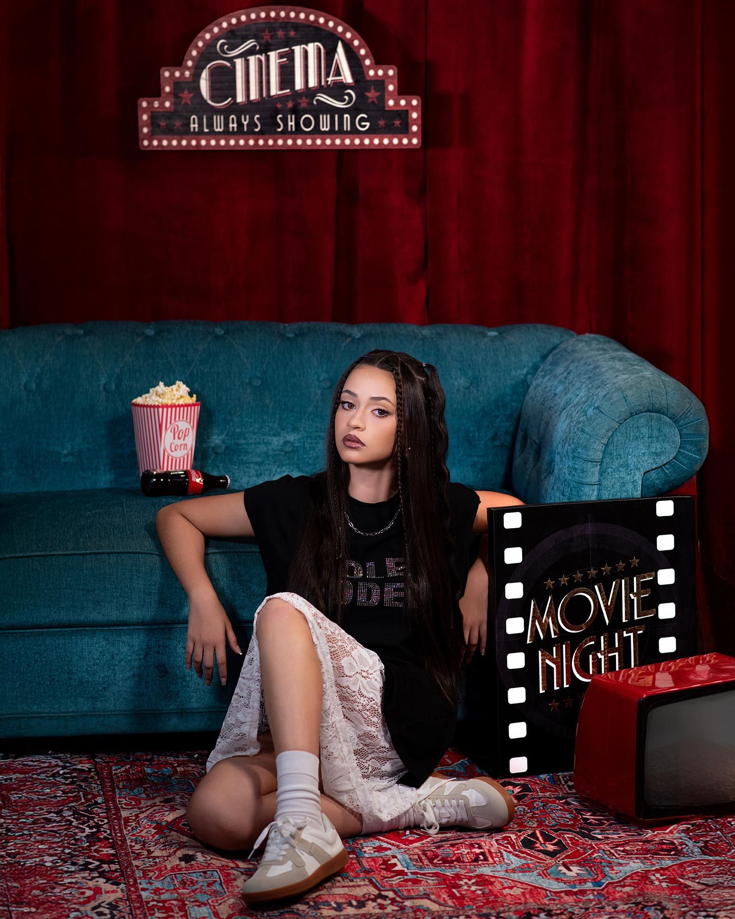 Movie night setup with vintage cinema sign, teal velvet couch, popcorn bucket and retro film decor on red carpet.