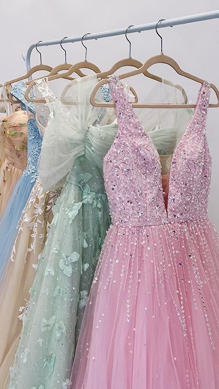Elegant evening gowns in pastel colors with sparkles and floral details hang on velvet hangers in a closet.