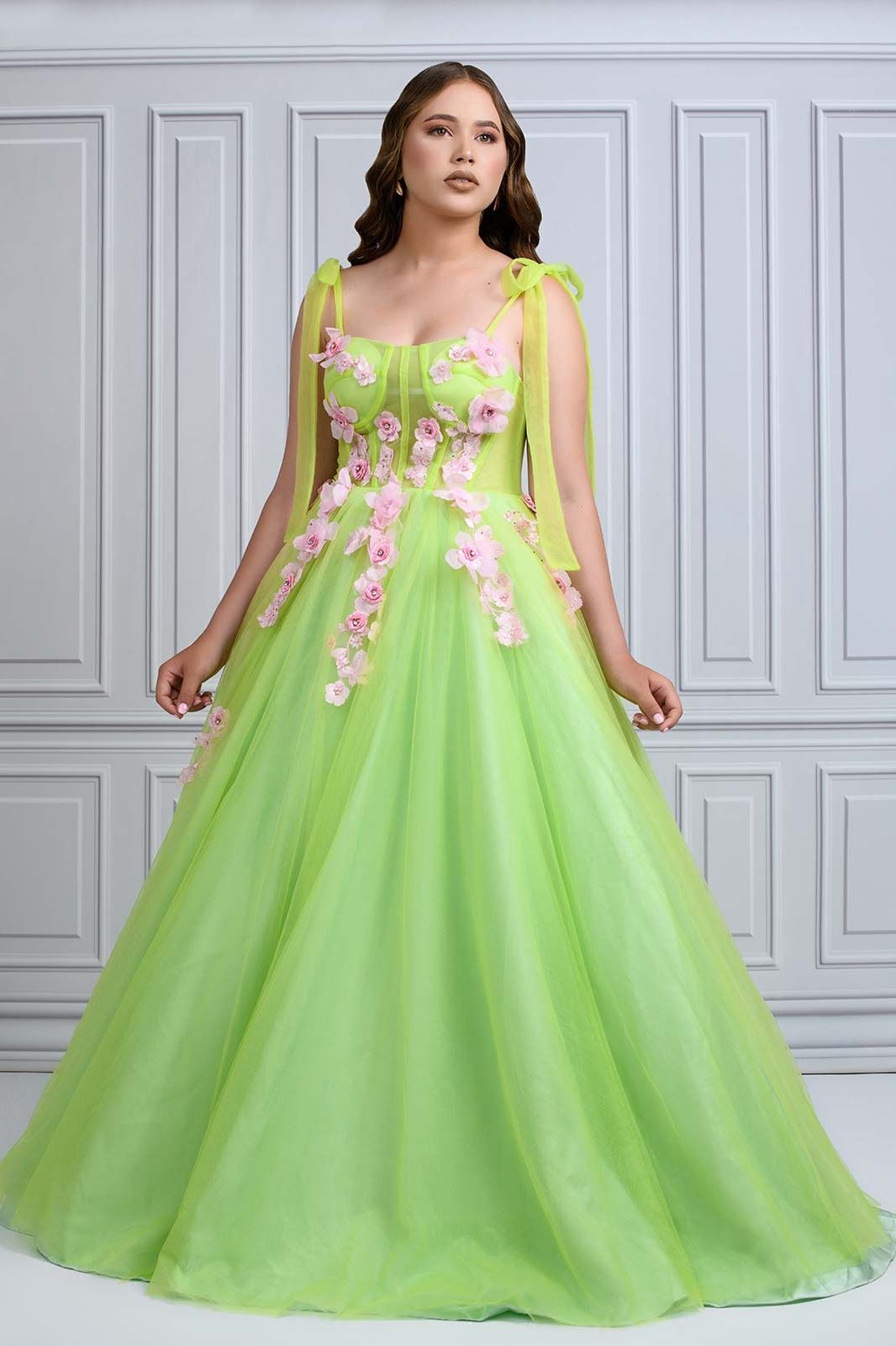 A lime green tulle ballgown with pink floral appliques and shoulder ties stands elegantly against a gray paneled wall.