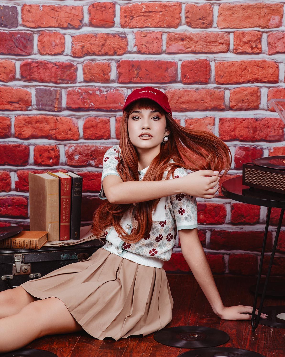 A vintage-styled fashion photo against a red brick wall featuring floral top and beige skirt with retro suitcases.