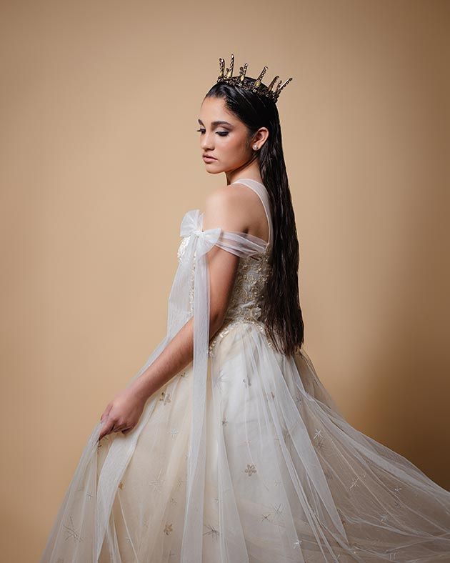 Elegant off-shoulder tulle ball gown with gold accents and delicate crown headpiece against beige backdrop.