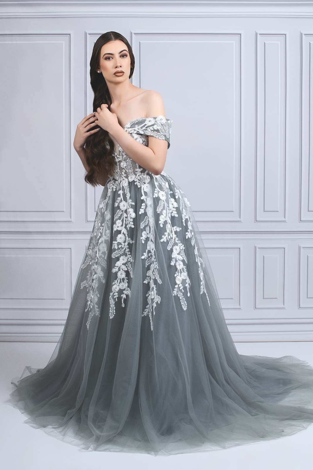An elegant gray tulle gown with white floral appliques and off-shoulder design against a classic wall.