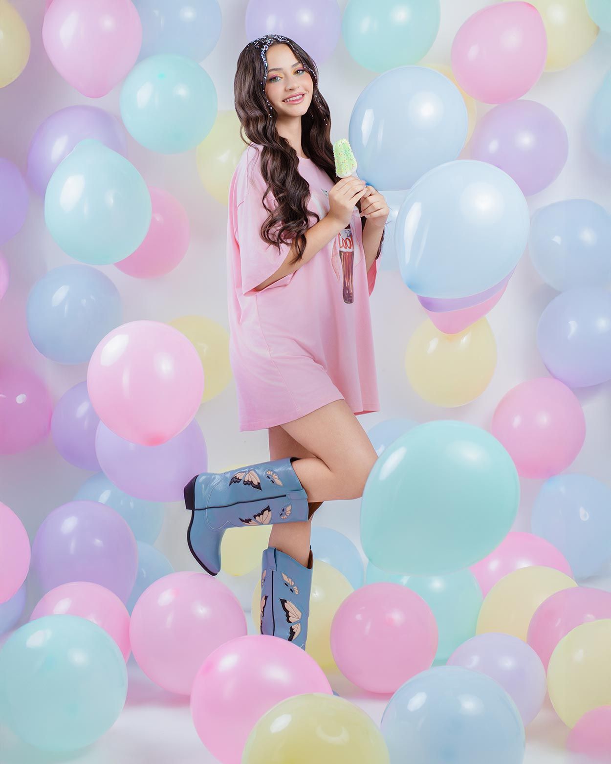 Model poses in pink dress and tall denim boots against pastel balloon backdrop with blue and pink balloons.