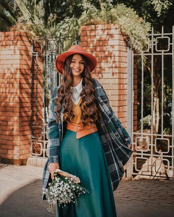 A vintage inspired fall outfit with plaid coat, rust hat, teal maxi skirt and small bouquet by brick pillars.