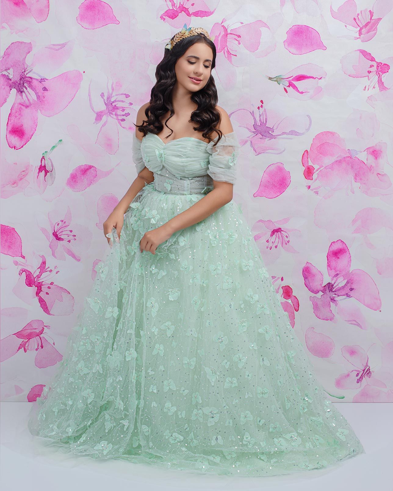 An elegant mint green ball gown with off-shoulder design and floral embellishments against a pink floral backdrop.