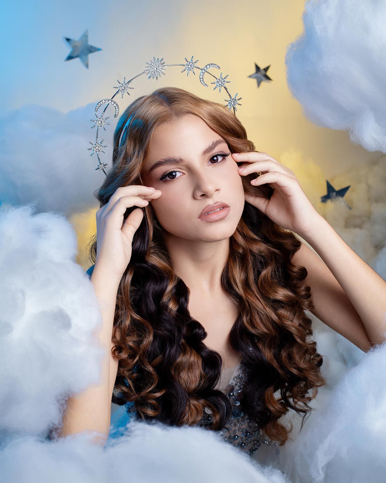 Ethereal portrait in clouds with stars showing long brown curls and a halo of snowflakes against blue and gold sky.