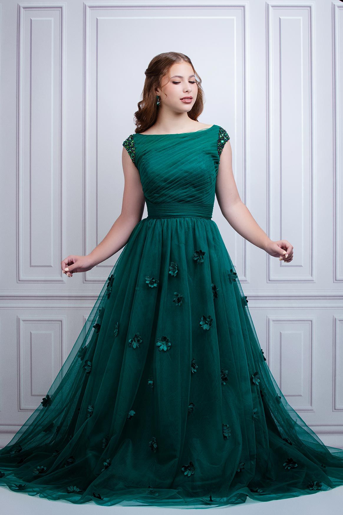 Elegant emerald green evening gown with delicate floral embellishments and boat neckline against white wall backdrop.