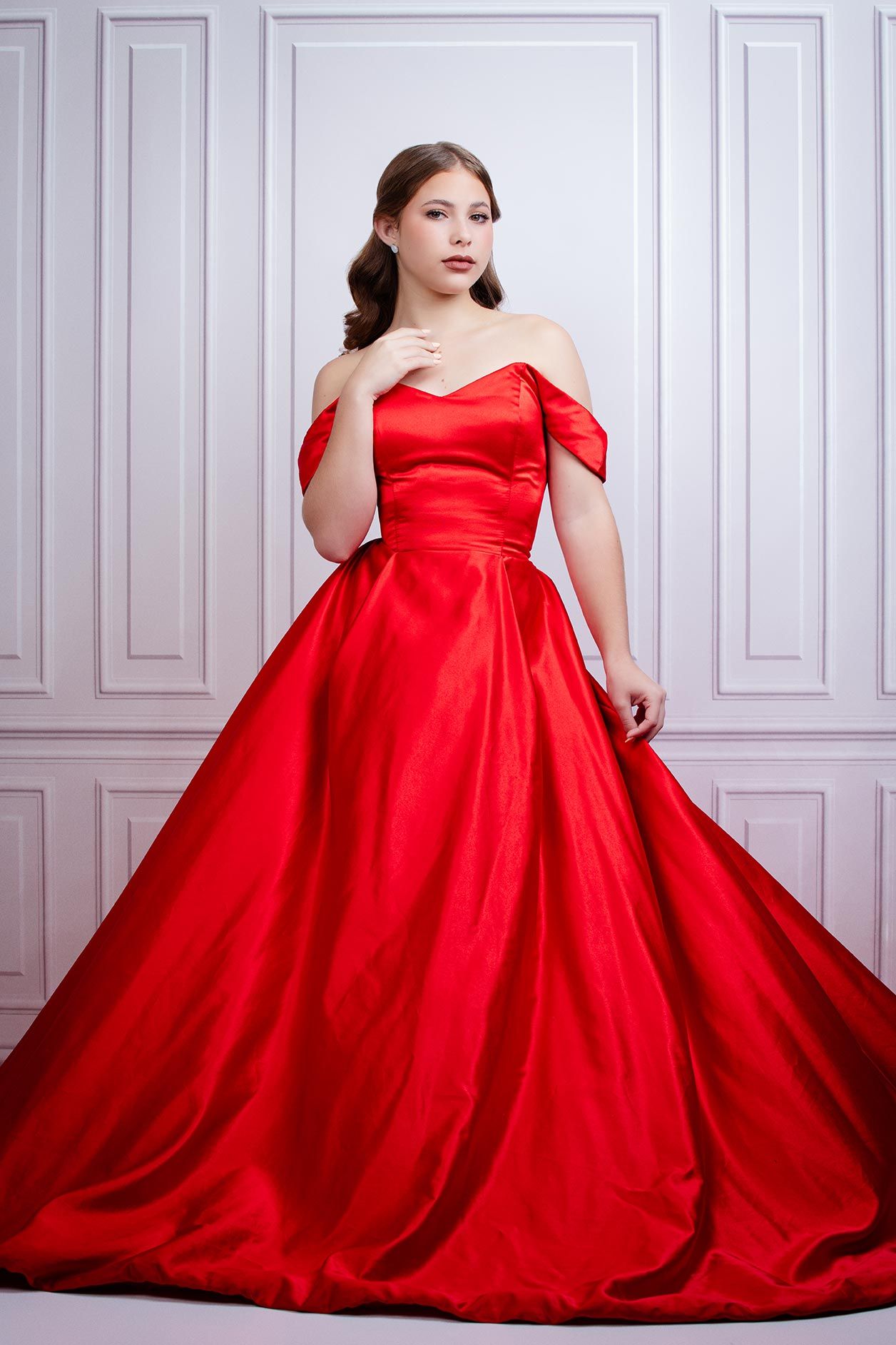A model poses in a glamorous red off-shoulder ball gown with full skirt against a white paneled wall.