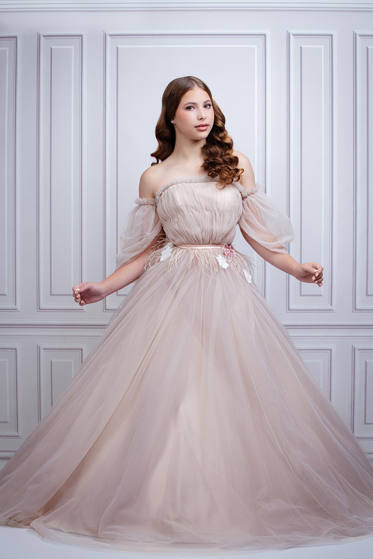A romantic off-shoulder blush tulle ball gown with puffy sleeves and delicate waist embellishments in a classic studio setting.