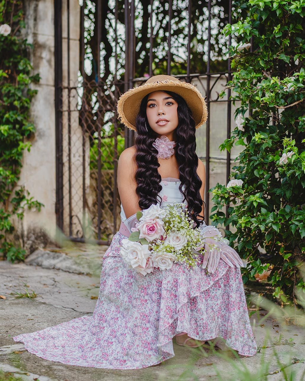 Woman in floral dress and straw hat seated with bouquet of white flowers in garden setting with climbing vines.