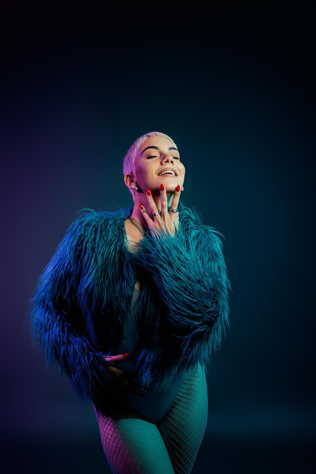 Artistic portrait in blue tones featuring a person in a fluffy teal feathered jacket posing dramatically against dark background.