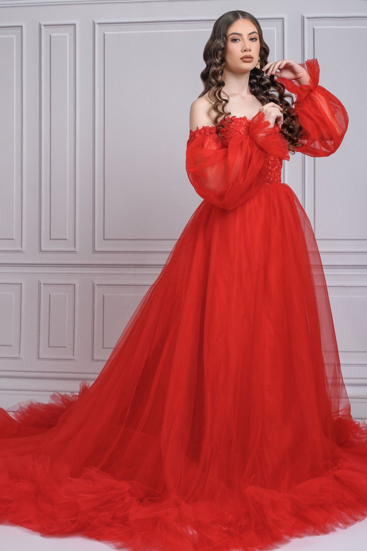 Dramatic red tulle ball gown with off-shoulder puffy sleeves and sweeping train against gray paneled wall.