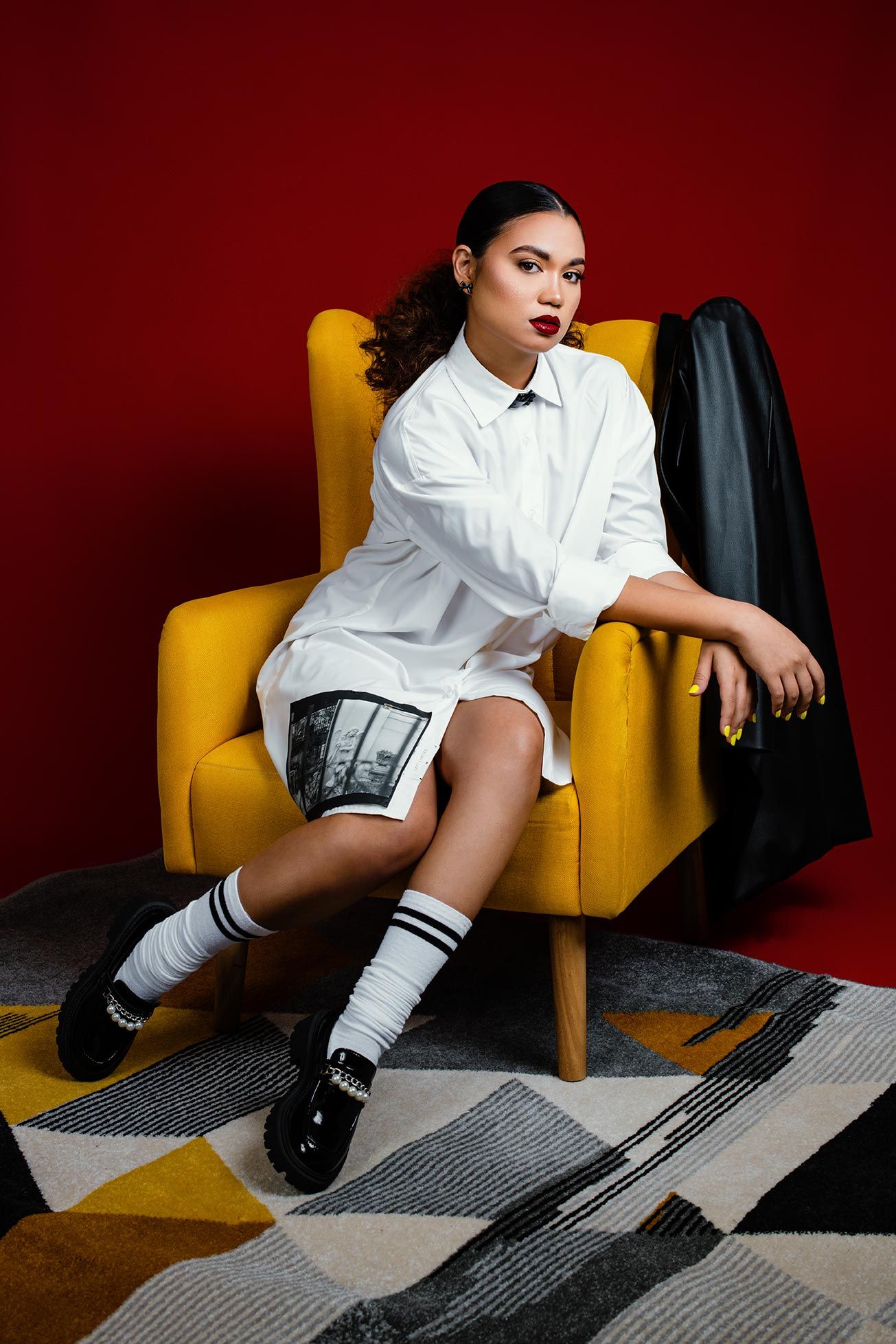 Stylish portrait against red wall showcasing white button-down shirt, striped socks and yellow armchair on geometric rug.