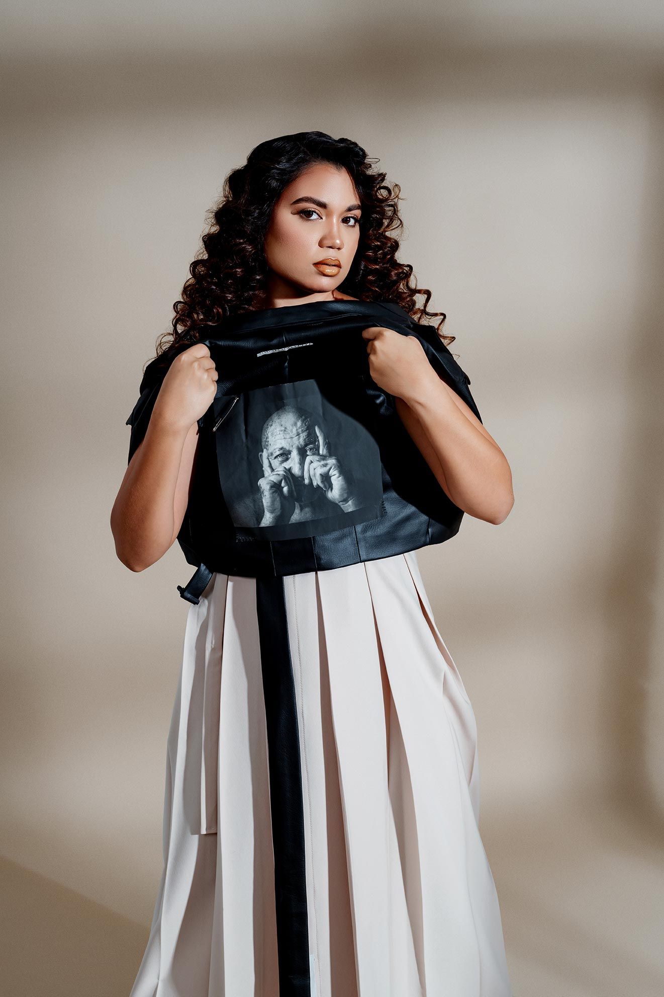 Fashion photo featuring dark curly hair against beige background wearing black top and white pleated skirt.