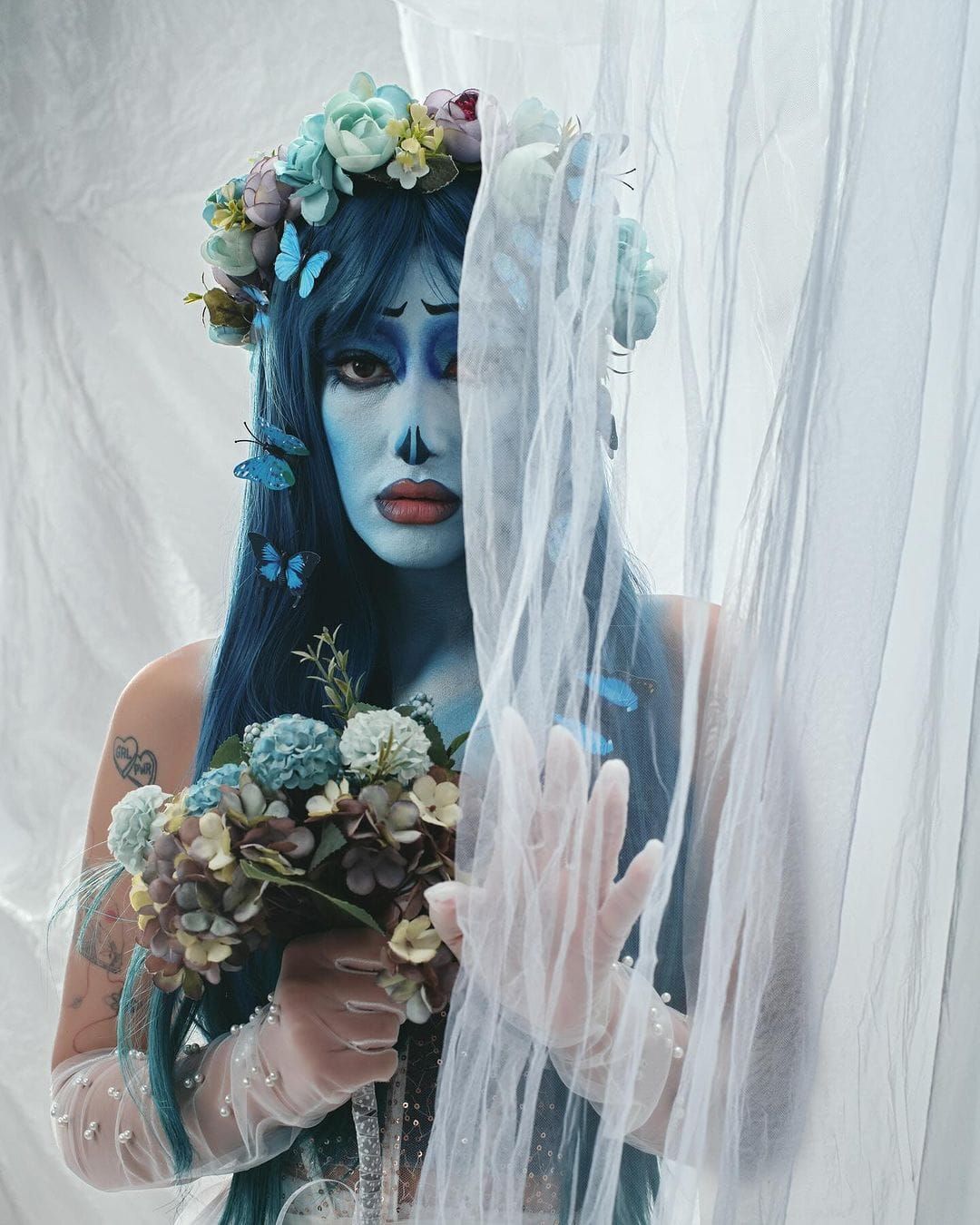 Fantasy portrait with blue body paint, flower crown, delicate white veil and floral decorations in an ethereal style.